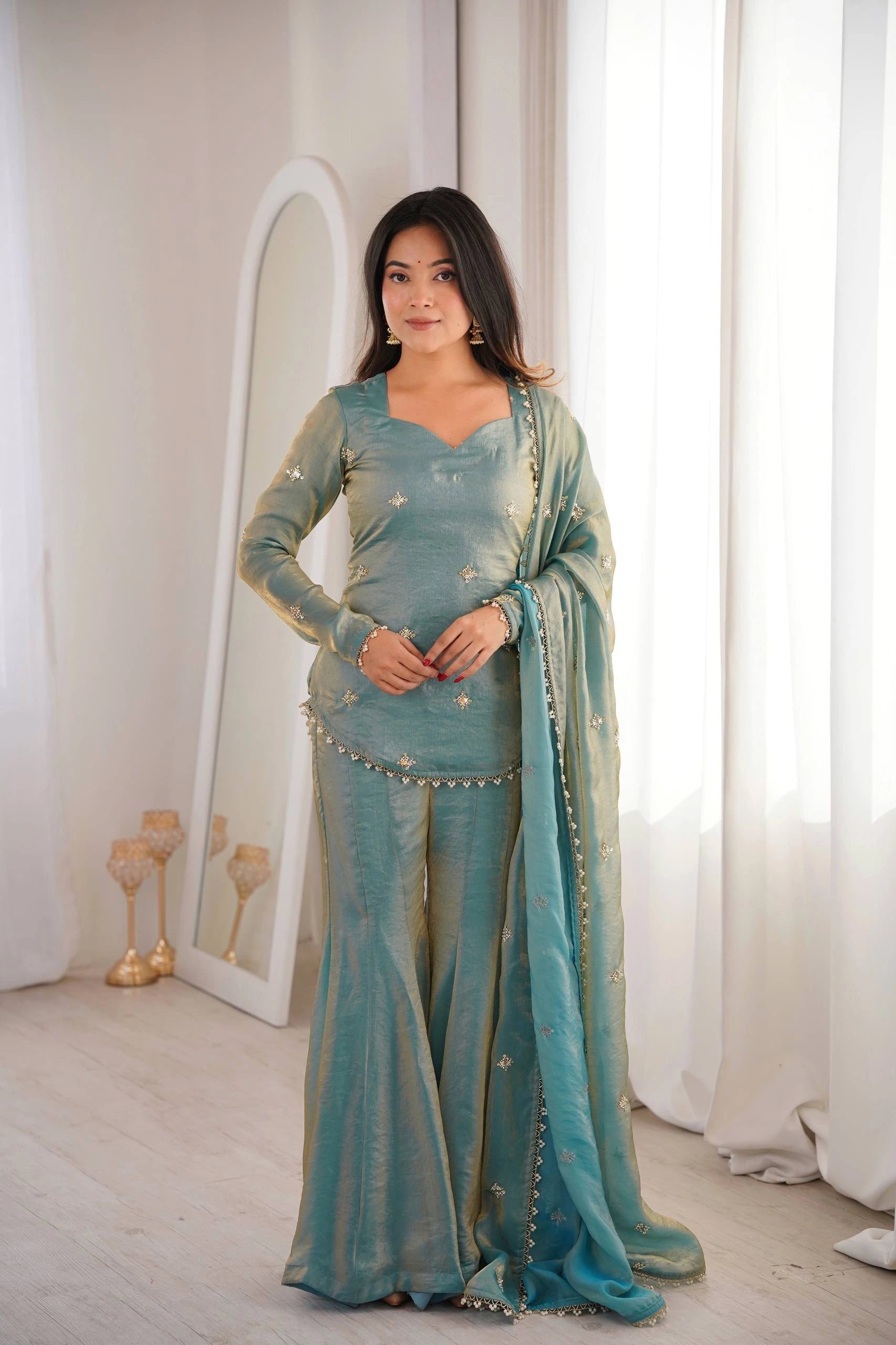 Woman in a teal traditional outfit standing in a room with white curtains and decor.